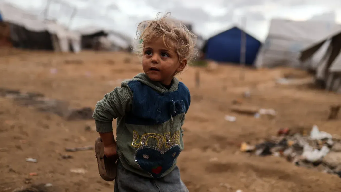 طفل صغير في مخيم للنازحين بغزة، يبدو عليه الخوف والقلق، وسط بيئة قاسية من الخيام والأنقاض، يعكس تأثير الحرب على الأطفال.