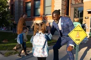 مدير سابق مبتسم يحيي طفلة صغيرة أمام مدرسة، بينما يظهر في الخلفية أشخاص آخرون ولافتة تحذيرية لعبور المشاة.