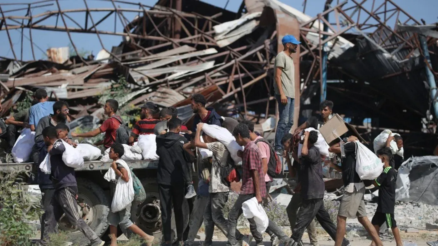 أطفال وراشدون يحملون مساعدات إنسانية في غزة، مع خلفية لمباني مدمرة، في سياق الأزمة الإنسانية المتفاقمة جراء الحرب.