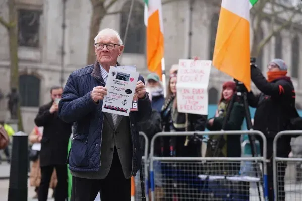رجل مسن يحمل لافتة خلال احتجاج في لندن، مع أعلام إيرلندية في الخلفية، تعبيرًا عن دعم لقضية جيري آدامز.