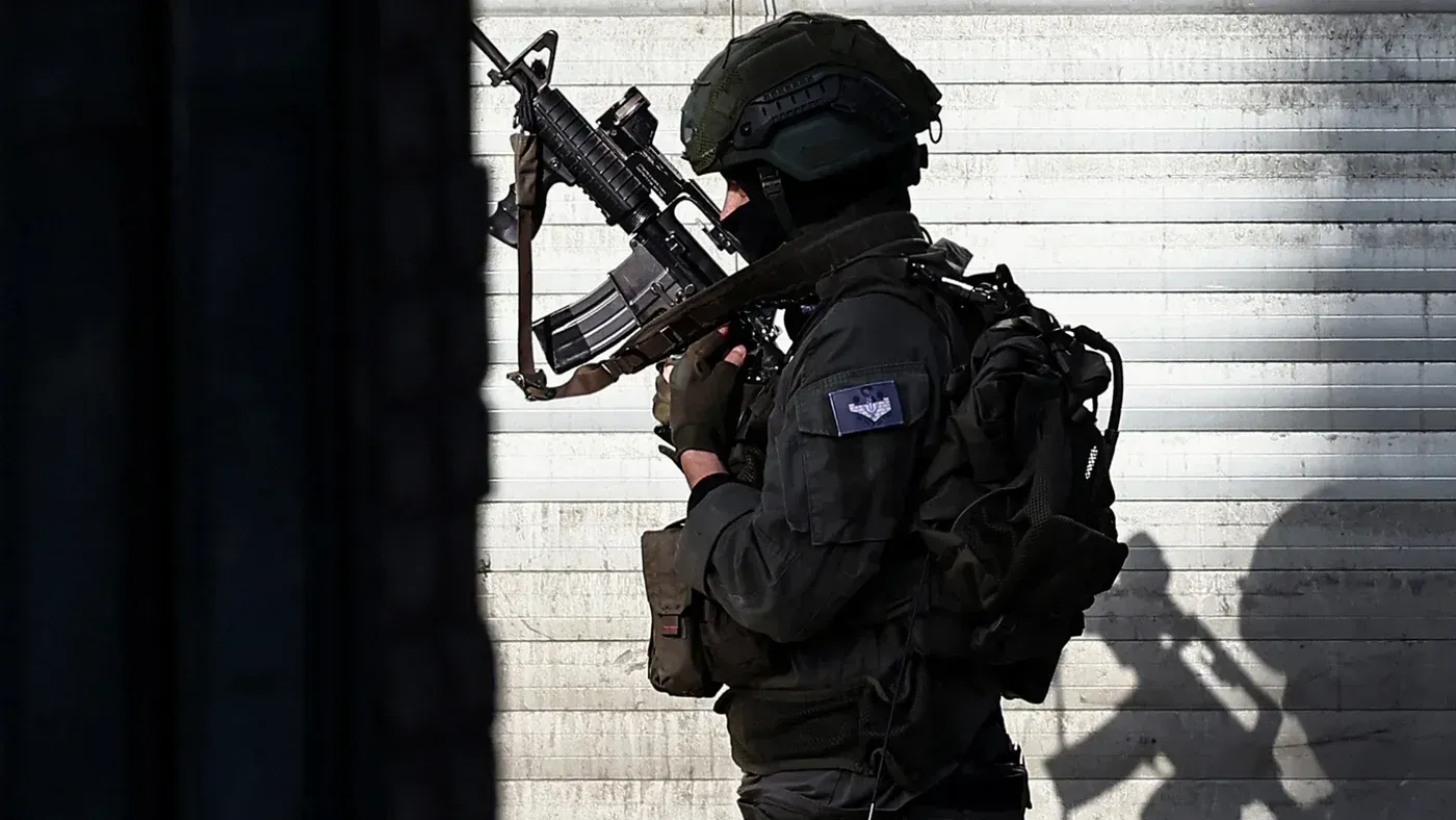 جندي إسرائيلي مسلح يقف بجانب جدار أثناء مداهمة في الضفة الغربية، مما يعكس التوترات المستمرة وتأثيرها على الفلسطينيين.