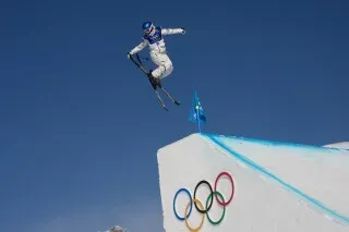 إيلين غو، متزلجة حرّة، تؤدي قفزة بهلوانية خلال التصفيات النهائية لسباق التزلج المنحدر في أولمبياد الشتاء، مع شعار الألعاب الأولمبية في الخلفية.