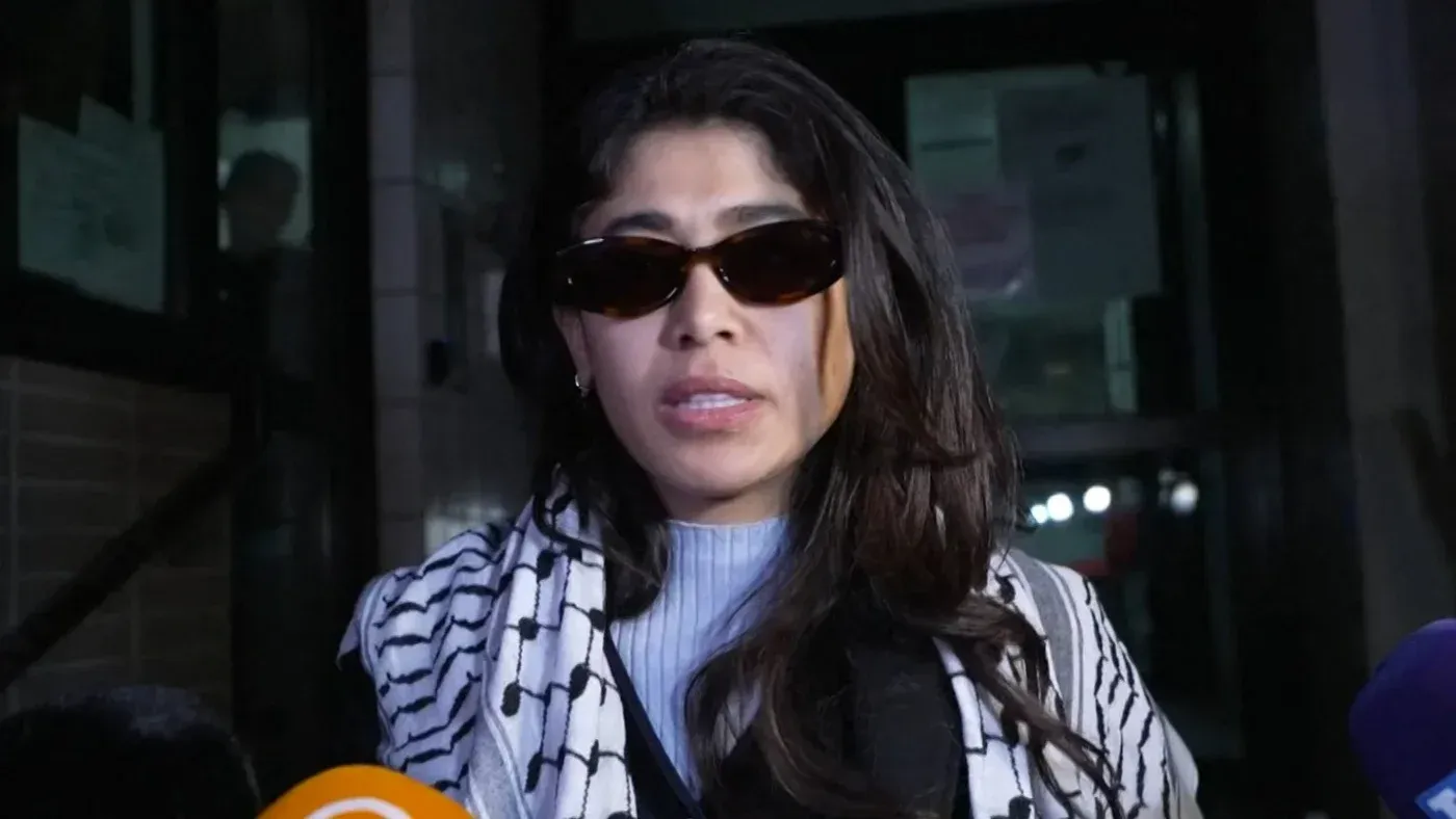 النائبة الفرنسية ريما حسن تتحدث للصحفيين بعد احتجازها، مرتدية نظارات شمسية، مع شال يحمل الألوان الفلسطينية.