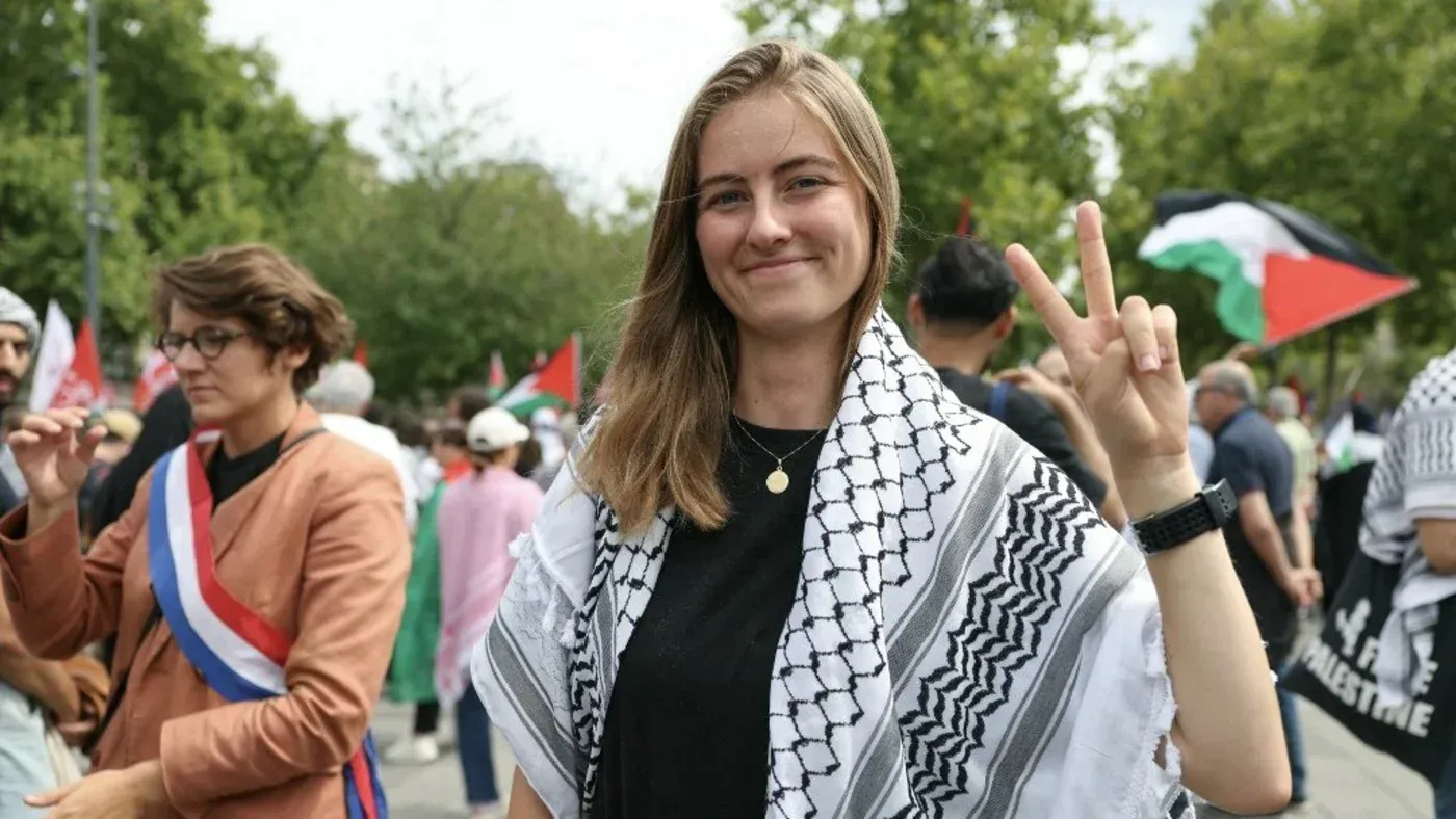 امرأة شابة ترتدي الكوفية الفلسطينية، تبتسم وتؤدي علامة النصر، وسط تجمع حاشد لدعم فلسطين.