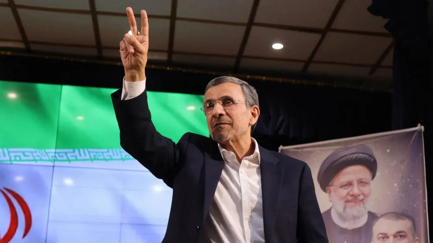 الرئيس الإيراني السابق محمود أحمدي نجاد يرفع إصبعيه في إشارة النصر، خلفه علم إيران، تعبيراً عن عودته المحتملة إلى الساحة السياسية.