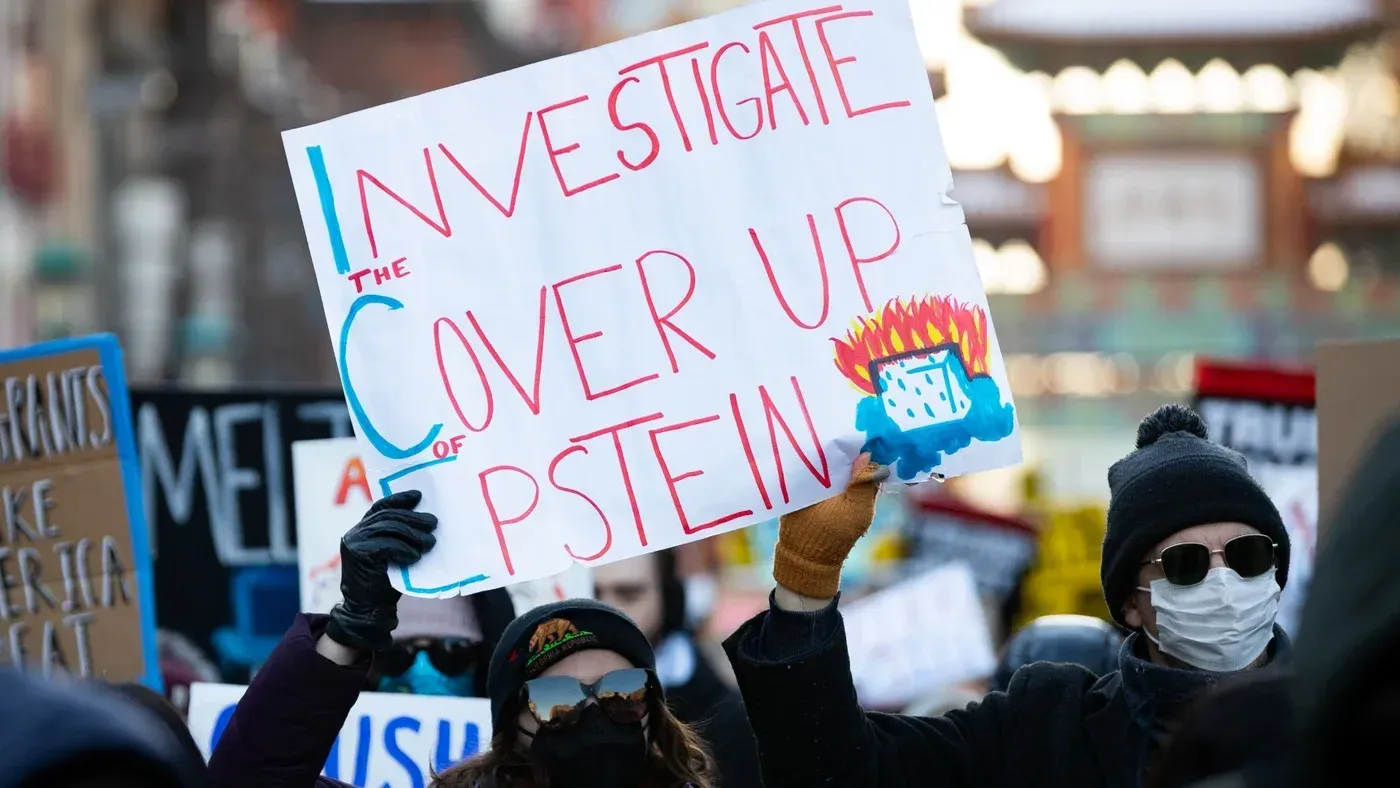 لافتة في مظاهرة تطالب بالتحقيق في قضية إبشتاين، مكتوب عليها "تحقيق في التستر على إبشتاين"، مع متظاهرين يرتدون أقنعة.
