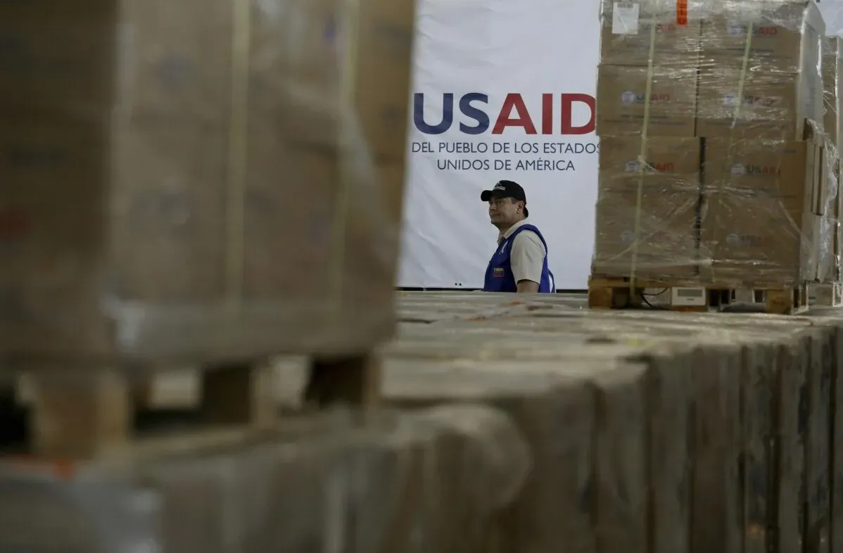 موظف في مركز توزيع المساعدات الإنسانية للوكالة الأمريكية للتنمية الدولية (USAID) بين صناديق الإغاثة، مع وجود لافتة خلفه توضح اسم الوكالة.