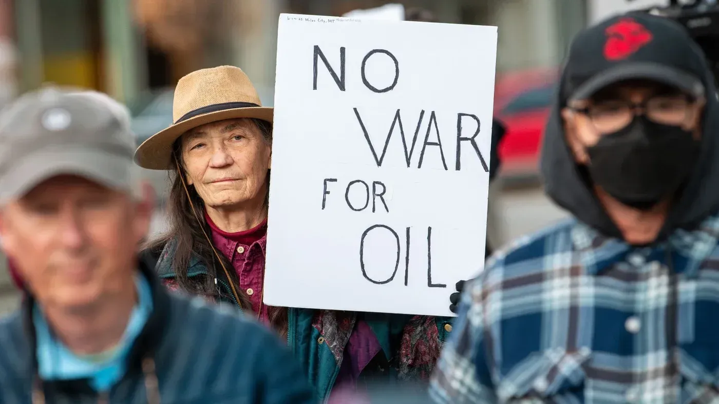 امرأة تحمل لافتة مكتوب عليها "لا للحرب من أجل النفط" في مظاهرة، بينما يظهر في الخلفية عدد من المتظاهرين. تعكس الصورة مشاعر المعارضة للحروب المرتبطة بالنفط.
