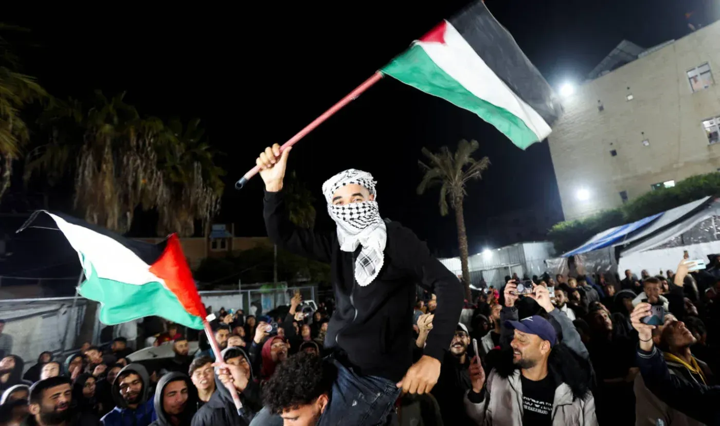 محتفل يحمل علم فلسطين وسط حشد من الناس في ليلة مظلمة، مع تواجد أشجار النخيل والمباني في الخلفية.