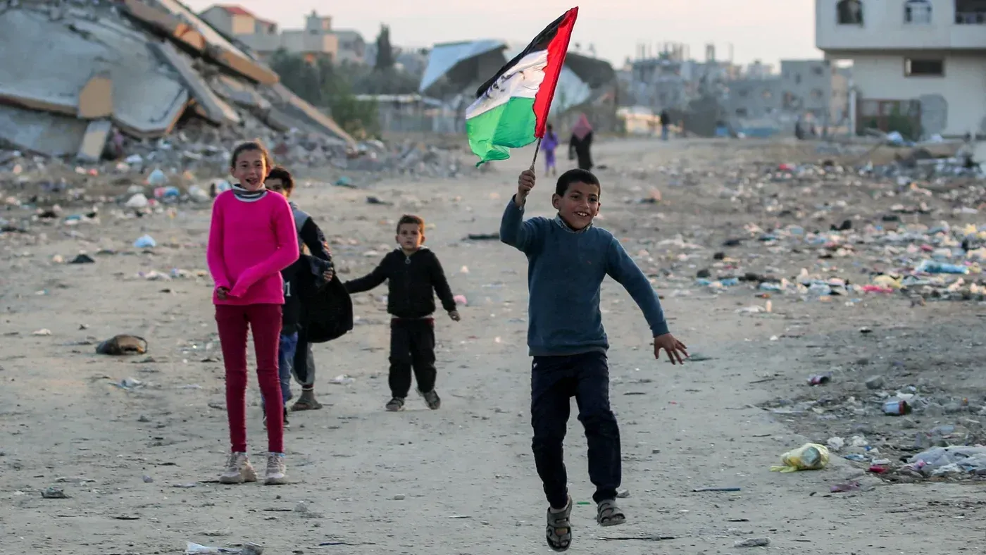أطفال فلسطينيون يلعبون في غزة، يحمل أحدهم علم فلسطين بفرح، خلفهم خلفية من الدمار والنفايات، تعبيرًا عن الأمل بعد وقف إطلاق النار.