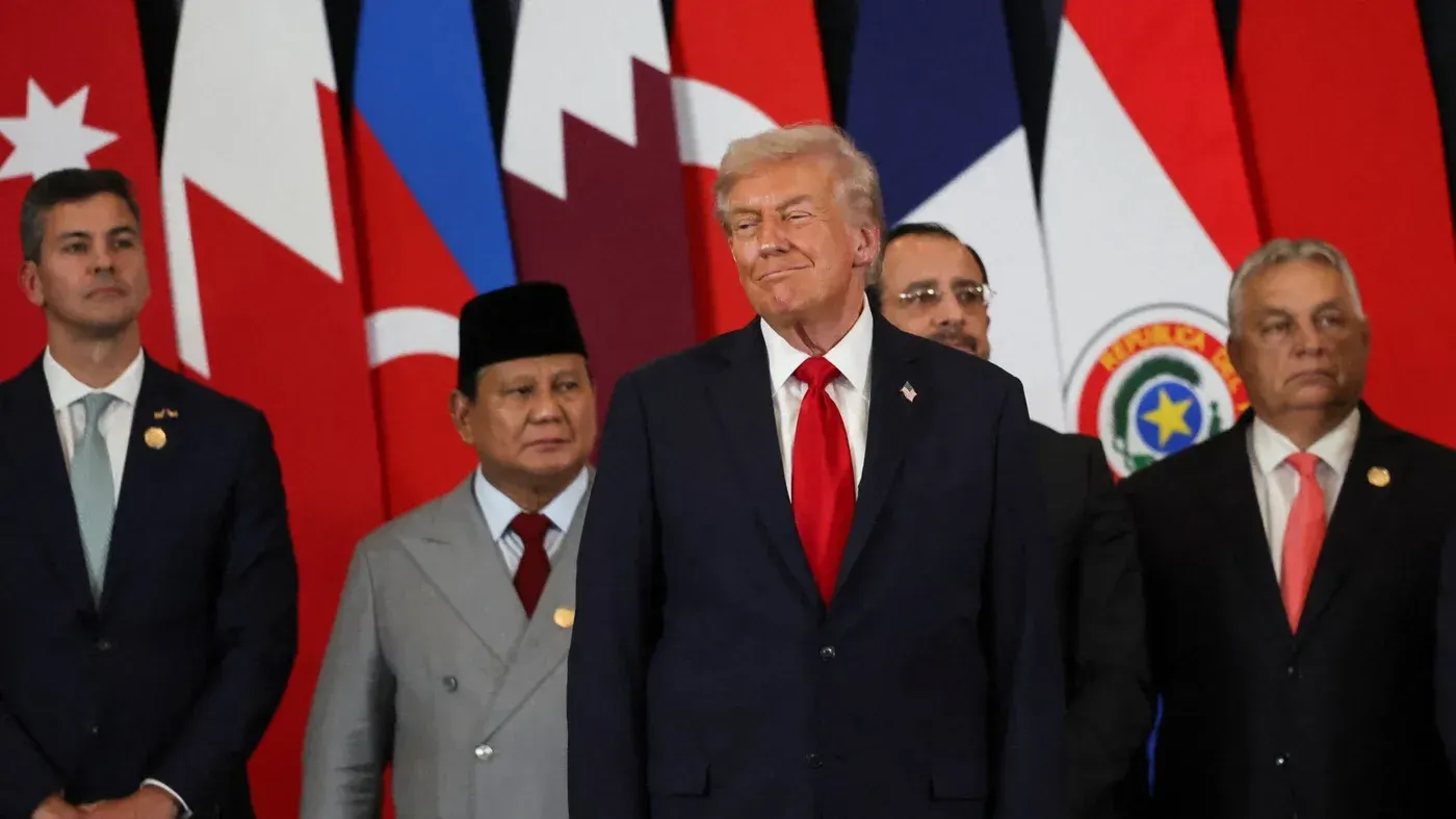 ترامب يقف مبتسمًا وسط قادة دوليين في قمة، مع أعلام متعددة خلفه، مما يعكس التوترات السياسية في الشرق الأوسط.