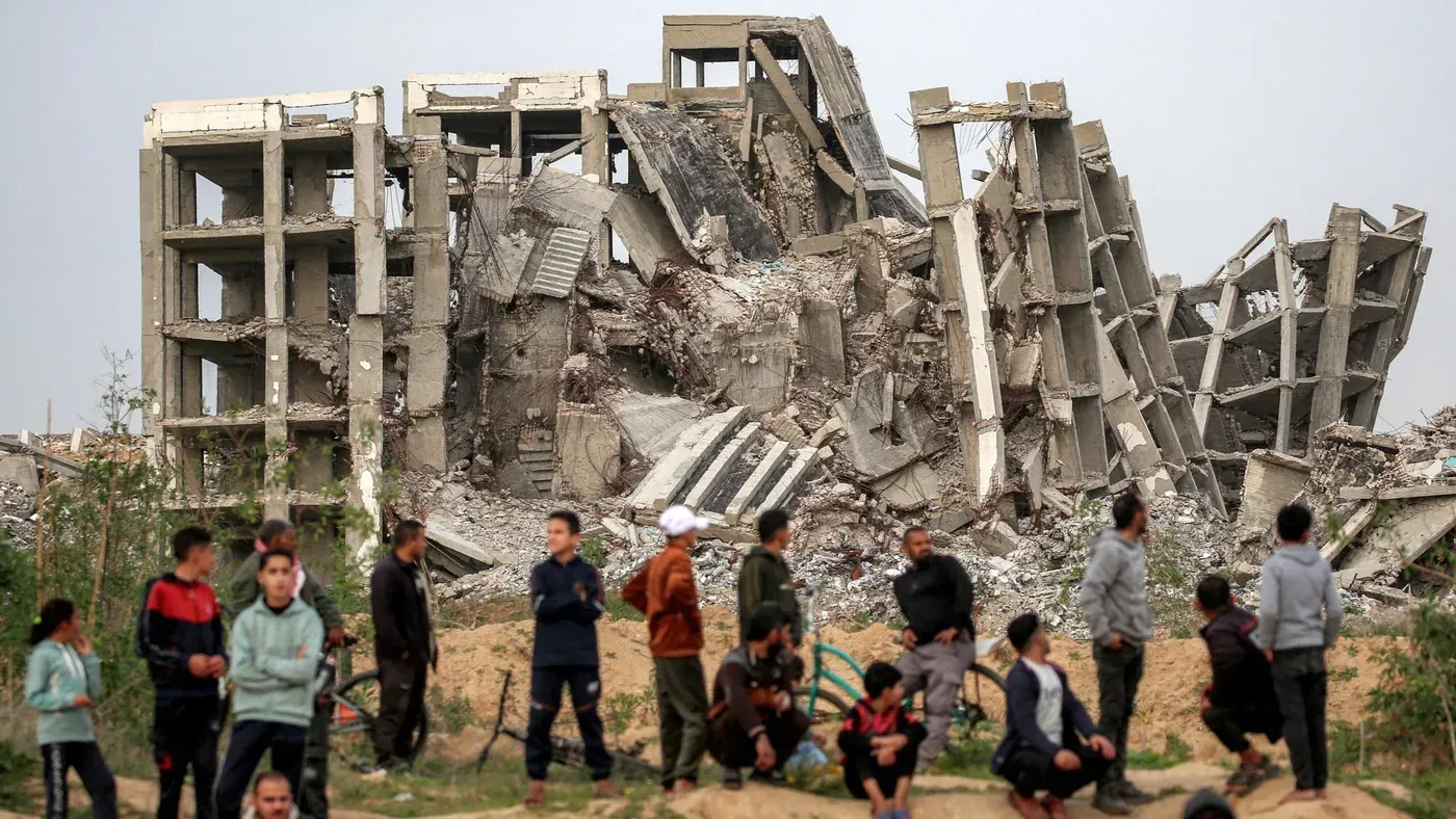 تظهر الصورة مجموعة من الأشخاص يقفون أمام مبنى مدمر في غزة، مما يعكس آثار الحرب والوفيات العنيفة التي تجاوزت 75,000 حالة.