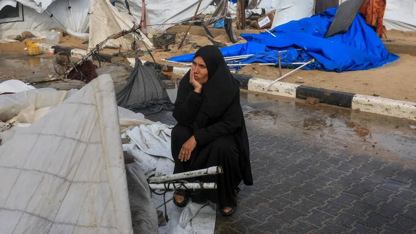 امرأة ترتدي الحجاب تجلس على الأرض وسط خيام مدمرة ومياه راكدة، تعكس معاناة سكان غزة خلال العواصف الشتوية.