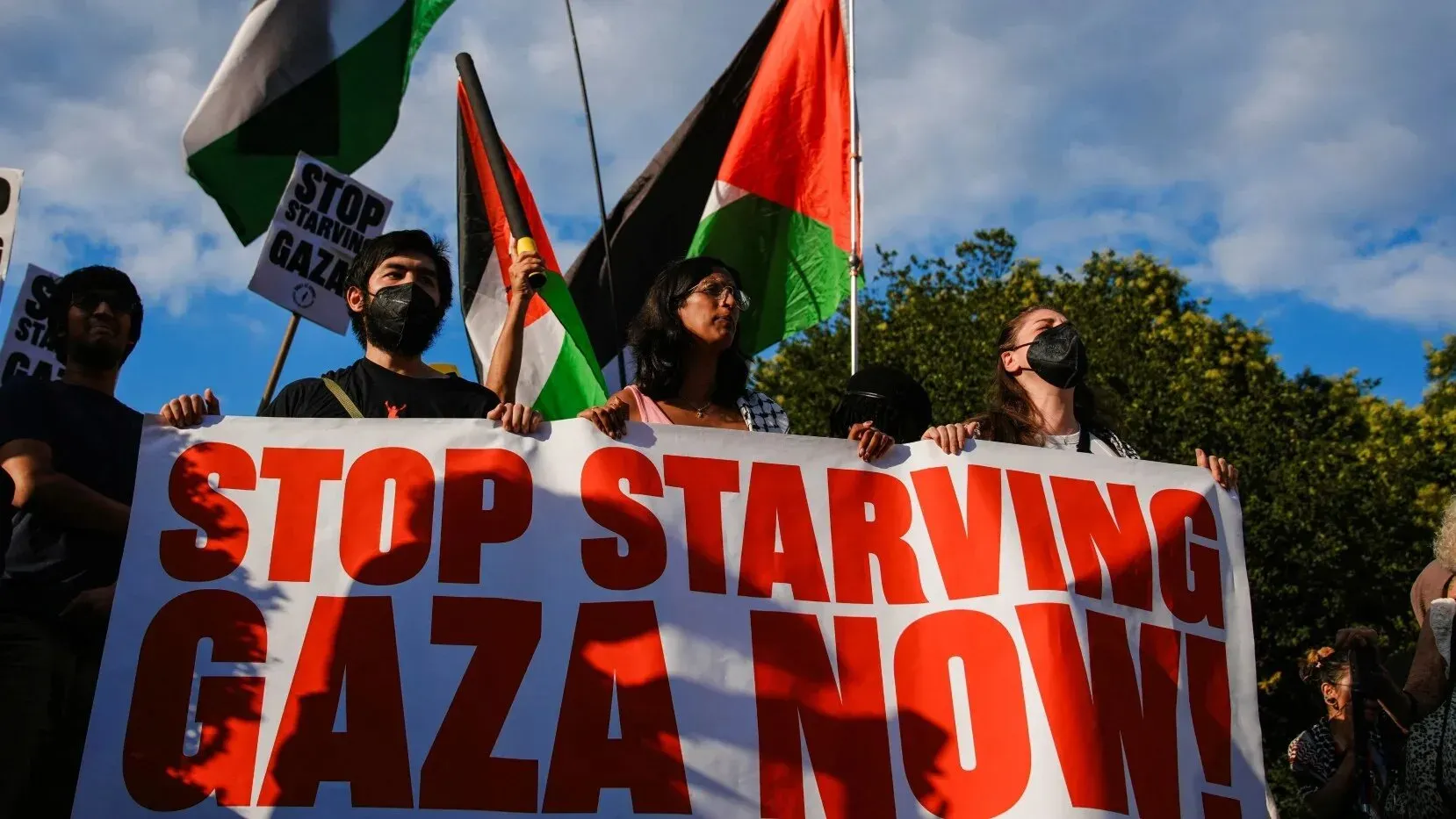 محتجون يحملون لافتة كبيرة مكتوب عليها "أوقفوا تجويع غزة الآن!"، مع أعلام فلسطينية في الخلفية، تعبيرًا عن الدعوة لإنهاء المجاعة.
