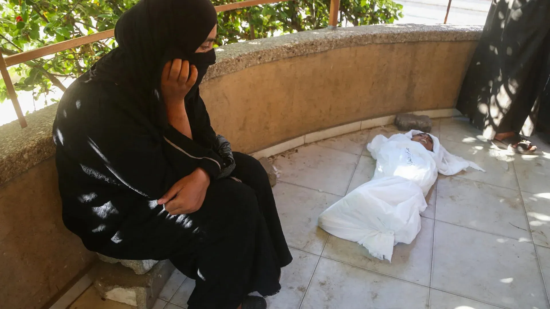 امرأة ترتدي الحجاب تجلس على حافة رصيف، تعبر عن الحزن بينما يستلقي طفل مغطى بغطاء أبيض على الأرض، في مشهد يبرز معاناة المجاعة في غزة.