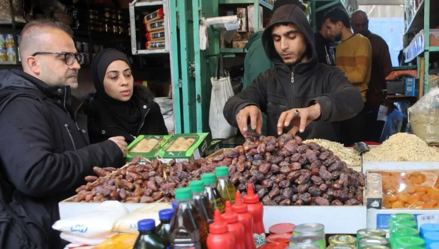 مشهد لثلاثة أشخاص يتسوقون في سوق غزة، حيث يظهرون أمام أكوام من التمر وزجاجات زيت، مع بائع يقوم بتحضير الطلبات.