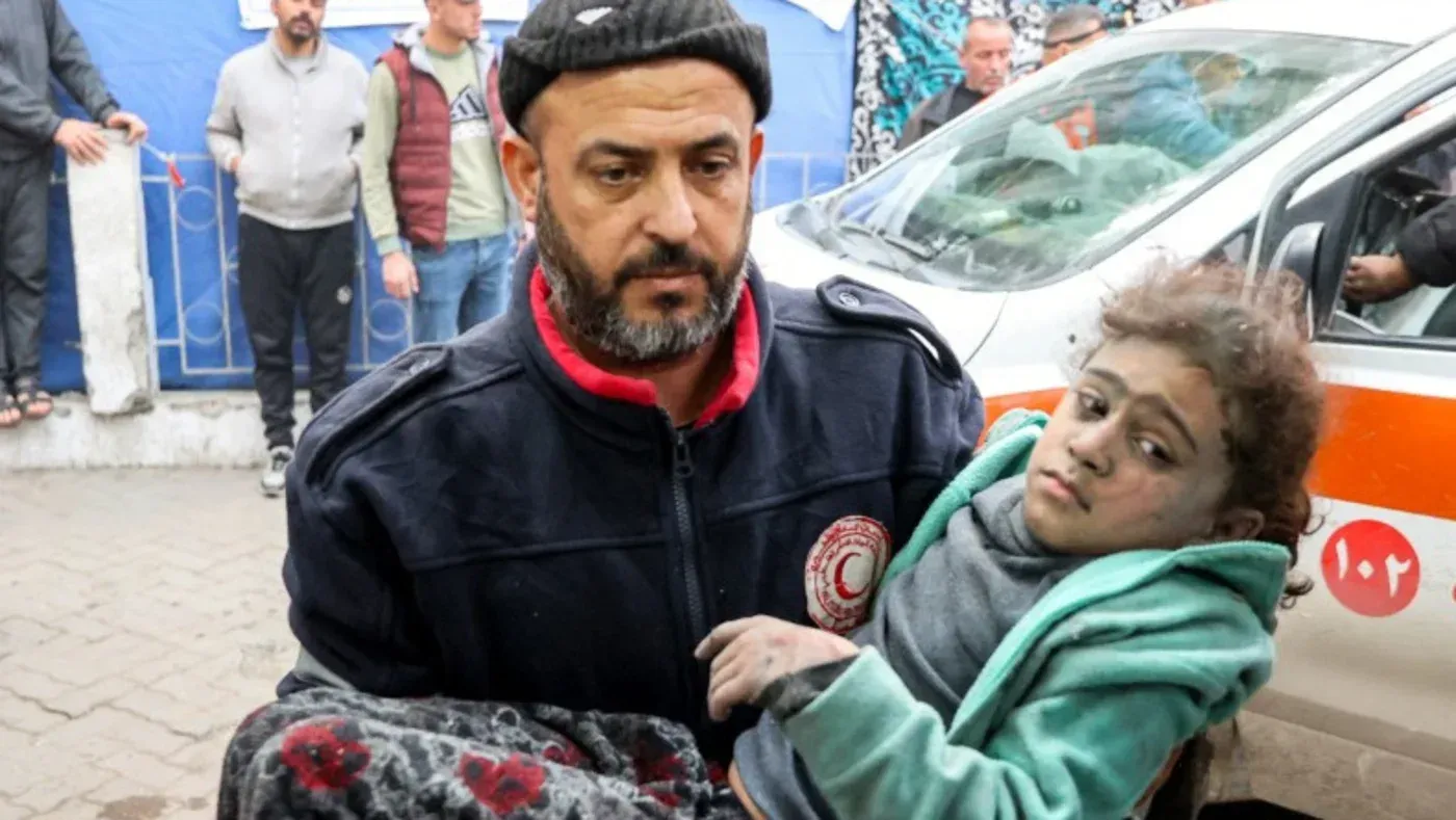 رجل يحمل طفلة مصابة، مغطاة بالتراب، وسط حشود في غزة، في ظل تصاعد الأوضاع الإنسانية بعد الهجمات الإسرائيلية.