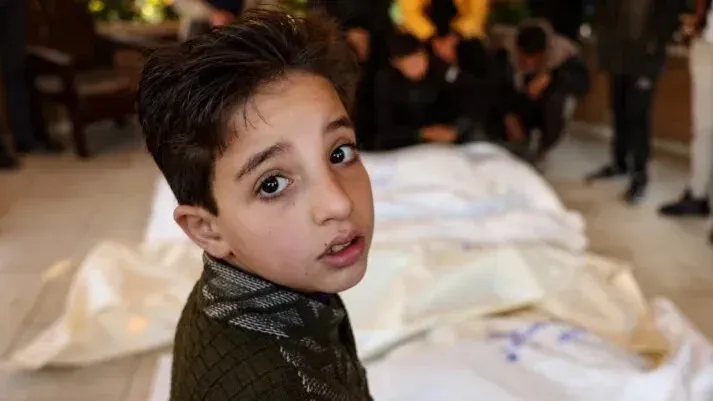 طفل فلسطيني ينظر بحزن إلى الكاميرا، محاط بجثث مغطاة، يعكس معاناة الأطفال في غزة خلال الإبادة الجماعية المستمرة.