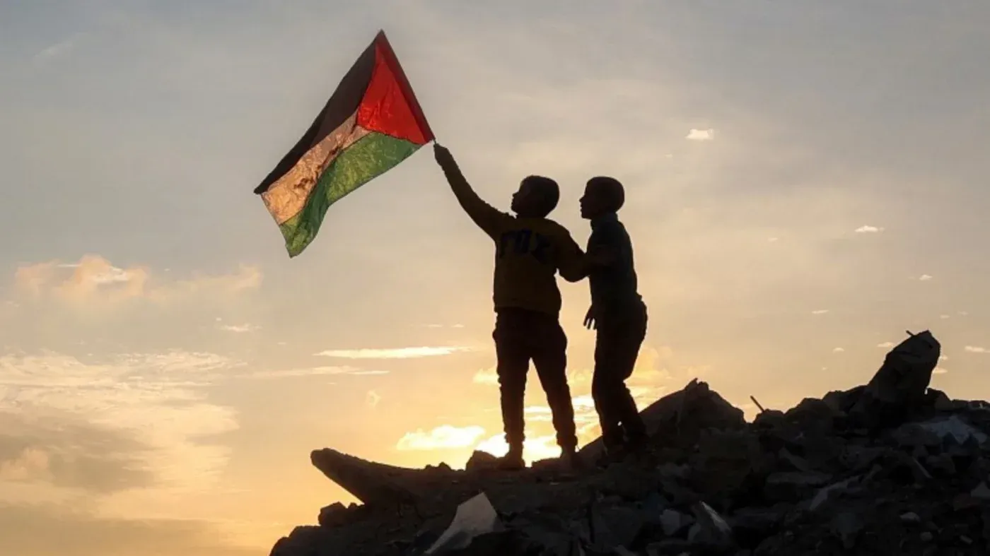 طفلان يحملان علم فلسطين في غروب الشمس، يقفان على أنقاض المباني المدمرة، معبرين عن الأمل في ظل الأوضاع الصعبة في غزة.