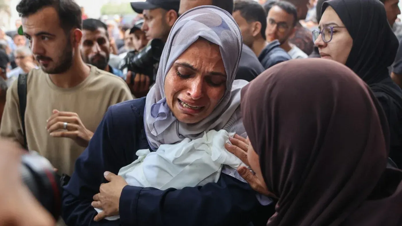 امرأة فلسطينية تبكي بينما تحمل طفلًا في حضنها، محاطة بجمهور متأثر، تعبيرًا عن الحزن والألم في ظل الظروف الصعبة في غزة.