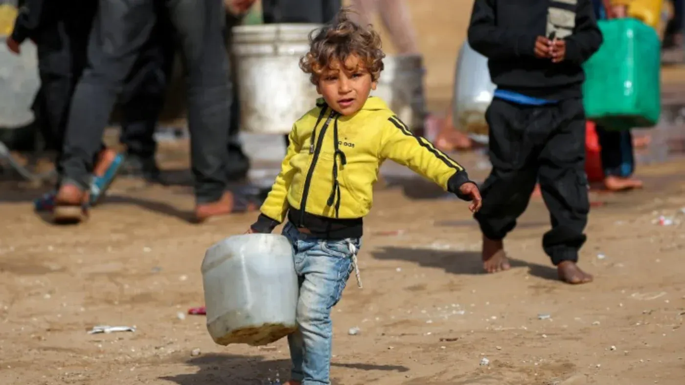 طفل صغير يحمل جرة مياه، يتنقل بين طوابير من الناس في غزة، مما يعكس أزمة المياه والاحتياجات الإنسانية المتزايدة في المنطقة.