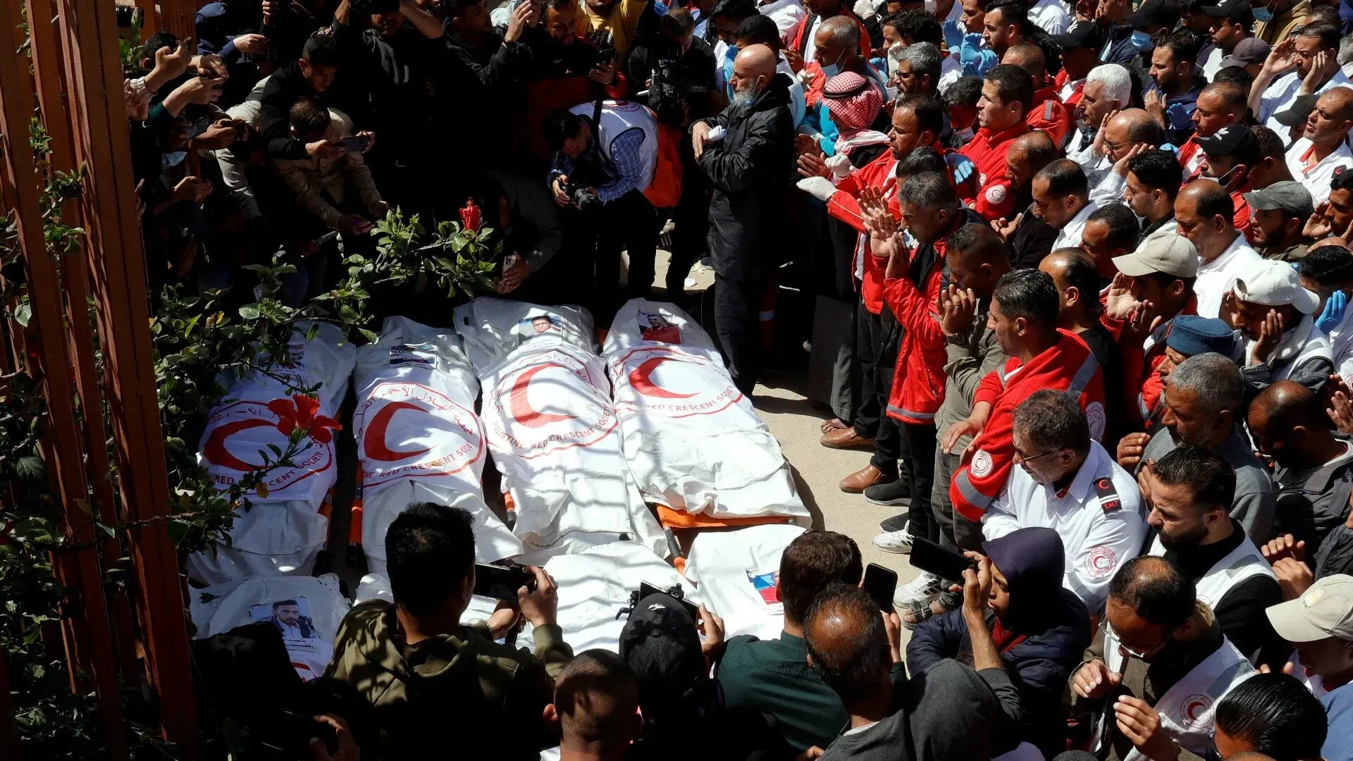 تجمع حشد كبير حول جثث مغطاة بأعلام جمعية الهلال الأحمر الفلسطيني، حيث يشارك الناس في مراسم تأبين لضحايا الهجمات الإسرائيلية.