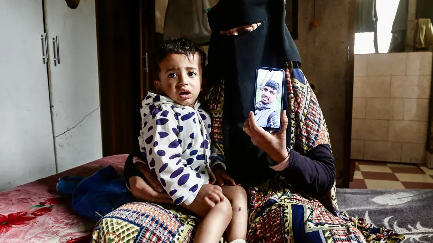 طفل فلسطيني يجلس في حضن والدته، يظهر على وجهه علامات الخوف، بينما تحمل والدته صورة لوالده.