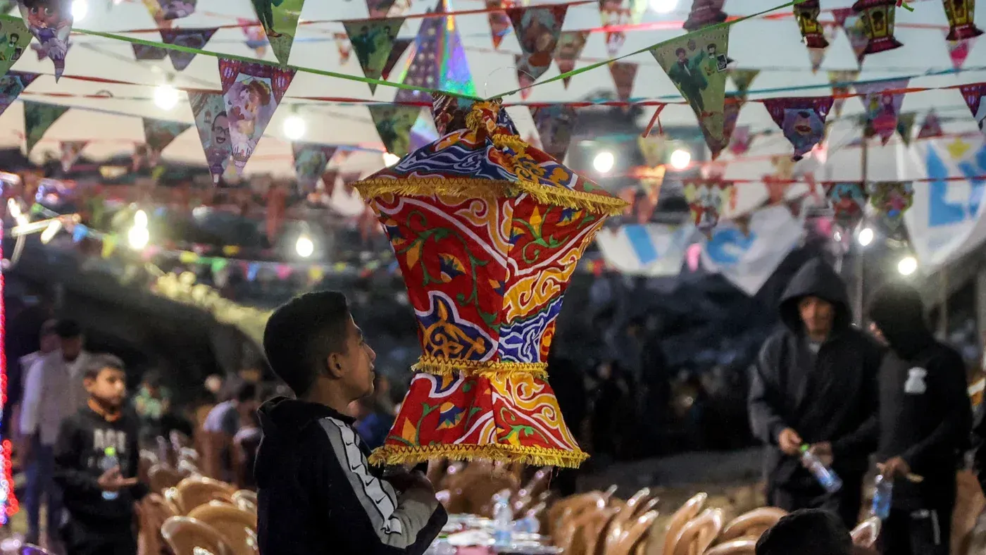 طفل يحمل فانوس رمضان ملون تحت زينة عيد رمضان في غزة، مع أضواء وزخارف تزين المكان، مما يعكس أجواء الاحتفال رغم الظروف الصعبة.