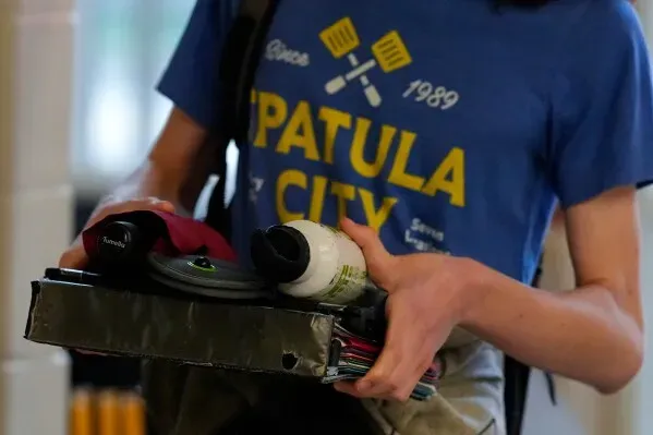 طالب يحمل مجموعة من الأدوات المدرسية في يده، يرتدي قميصًا مكتوبًا عليه "مدينة السباتولا" في سياق مناقشة حول فحص الأسلحة في المدارس.