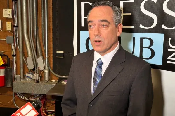 رجل يرتدي بدلة رسمية يتحدث في مؤتمر صحفي، مع خلفية تحمل شعار نادي الصحافة. يناقش قضايا انتخابية تتعلق بفواتير الكهرباء في جورجيا.