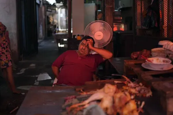 رجل يجلس في سوق، يبدو متعبًا ويضع يده على رأسه، بينما يعمل مروحة في الخلفية، مما يعكس تأثير درجات الحرارة المرتفعة.