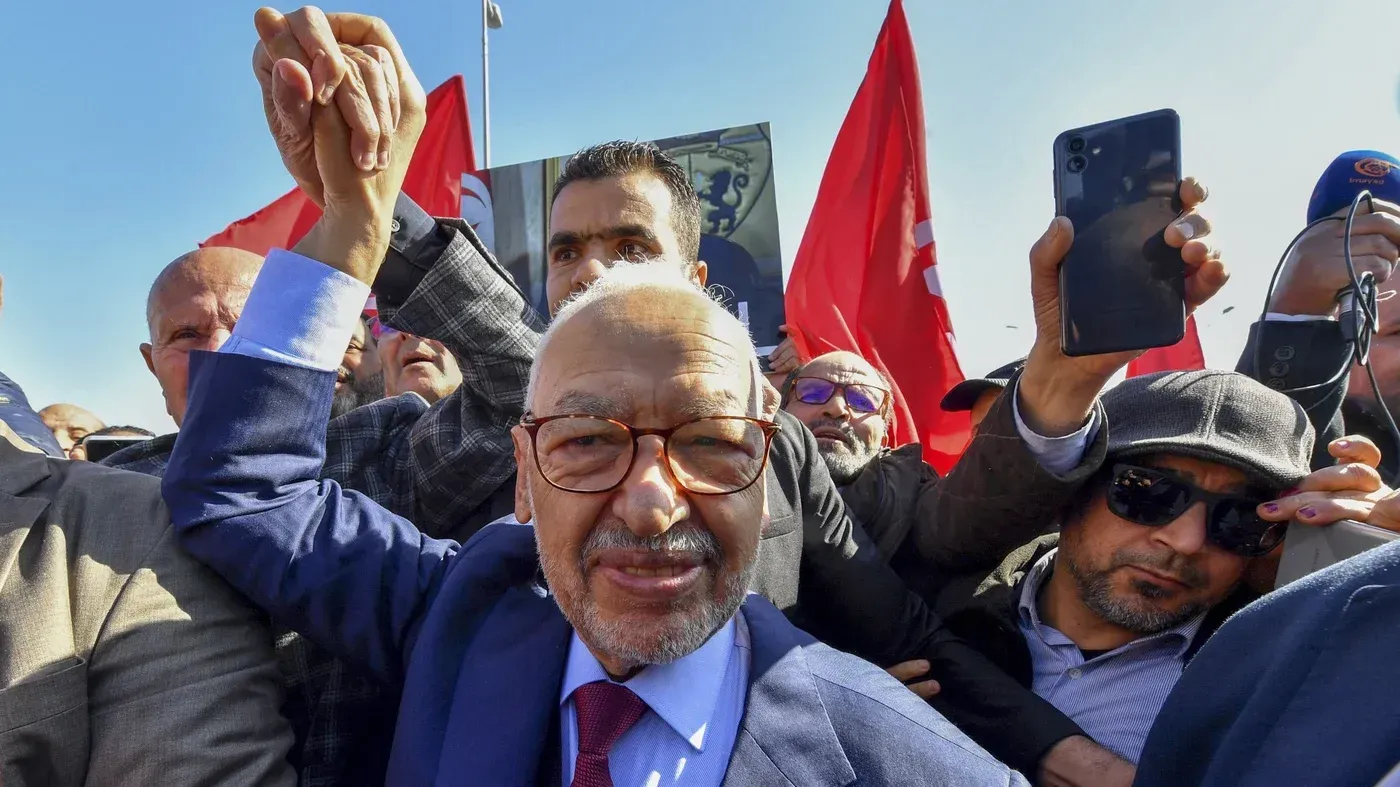 راشد الغنوشي، زعيم حزب النهضة، يبتسم ويرفع يده وسط حشد من المؤيدين، مع لافتات حمراء في الخلفية، في سياق الاحتجاجات السياسية في تونس.