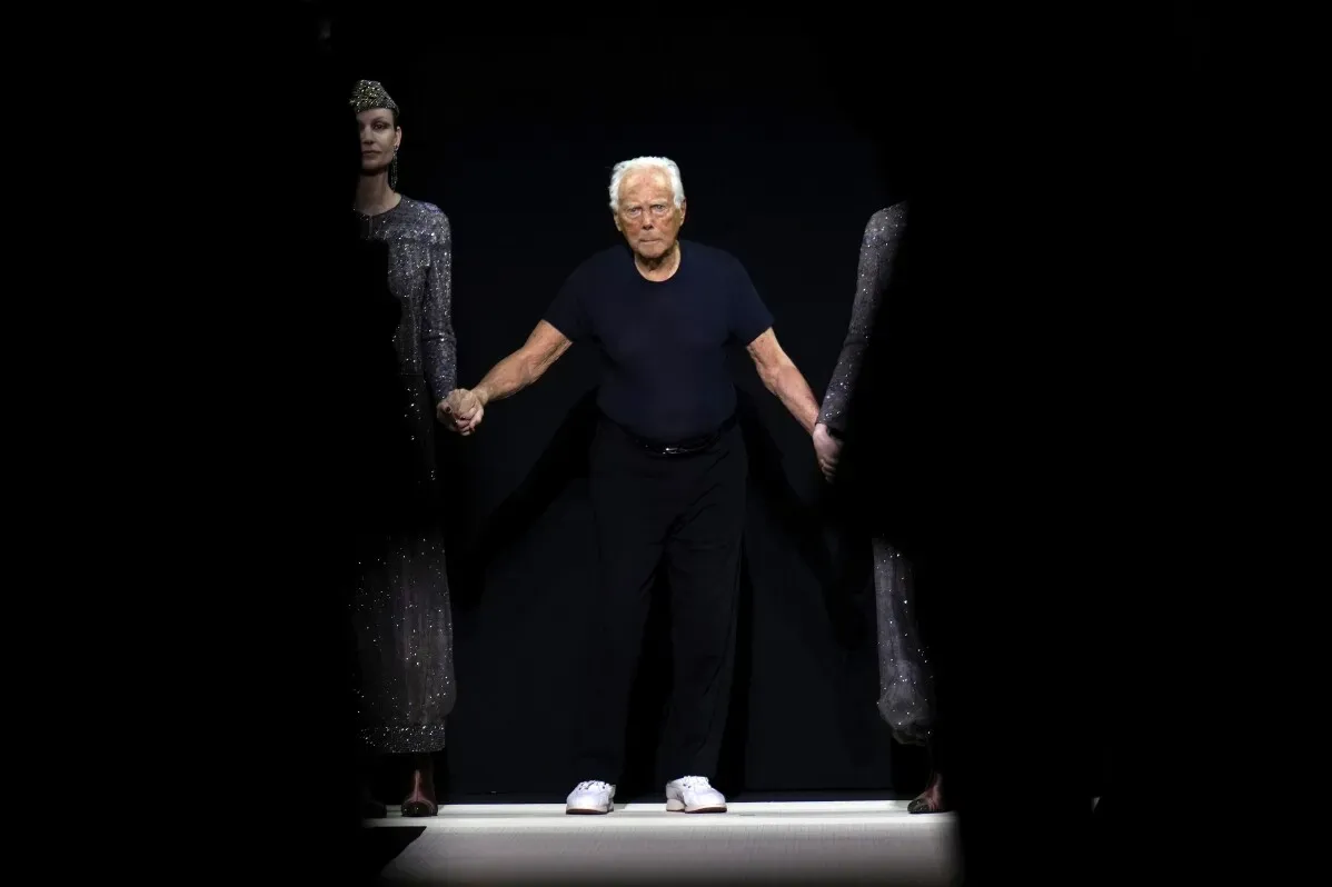 جورجيو أرماني يتوسط عارضتين أثناء انحنائه الختامي في عرض أزياء خريف وشتاء 2025-2026 في ميلانو، مع تسليط الضوء على الأناقة والحرفية.