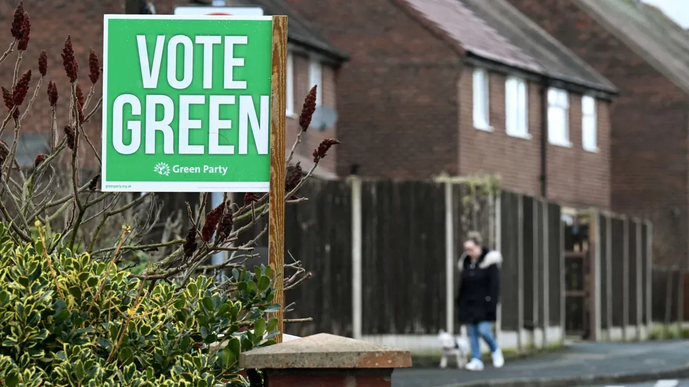 لافتة انتخابية لحزب الخضر مكتوب عليها "صوتوا للخضر"، مع مشاة في الخلفية يتجولون في حي سكني في مانشستر.