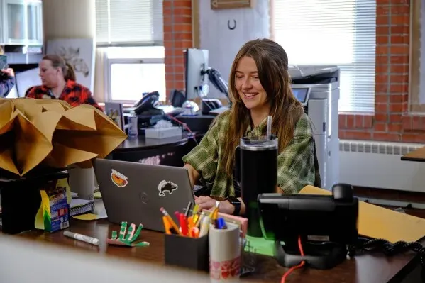 طالبة جامعية تعمل على الكمبيوتر في مكتب مدرسة مورنينغسايد الابتدائية، محاطة بأدوات مكتبية وأوراق، تعكس بيئة تعليمية نشطة.