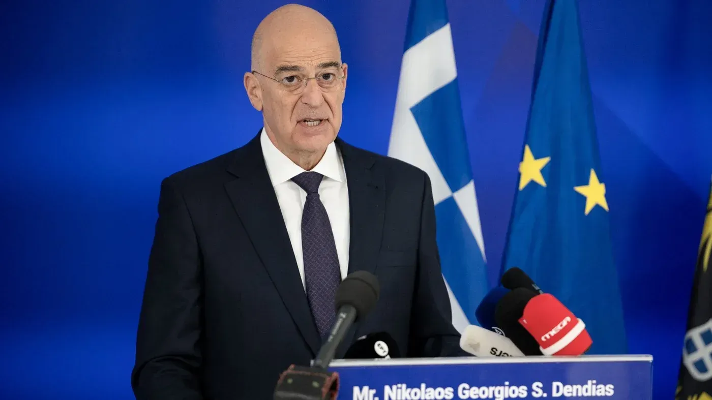 وزير الدفاع اليوناني نيكوس دندياس يتحدث في مؤتمر صحفي، مع وجود أعلام اليونان والاتحاد الأوروبي خلفه، مع التركيز على توتر العلاقات مع تركيا.