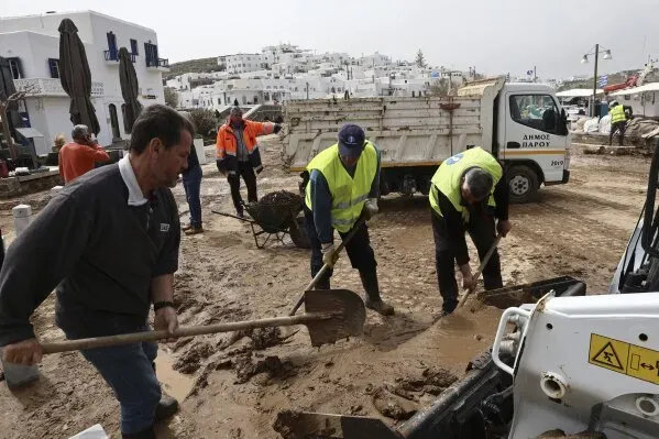 عمال يقومون بتنظيف الشوارع المليئة بالطين بعد الفيضانات في باروس، اليونان، مع وجود معدات ثقيلة في الخلفية.