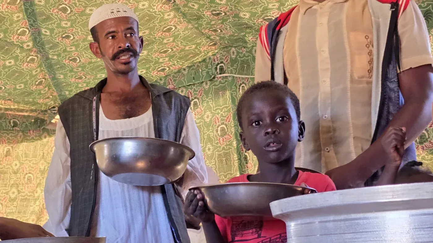 رجل وطفل يحملان أواني طعام في مخيم للاجئين بالسودان، وسط أزمة إنسانية تتعلق بالجوع وسوء التغذية.