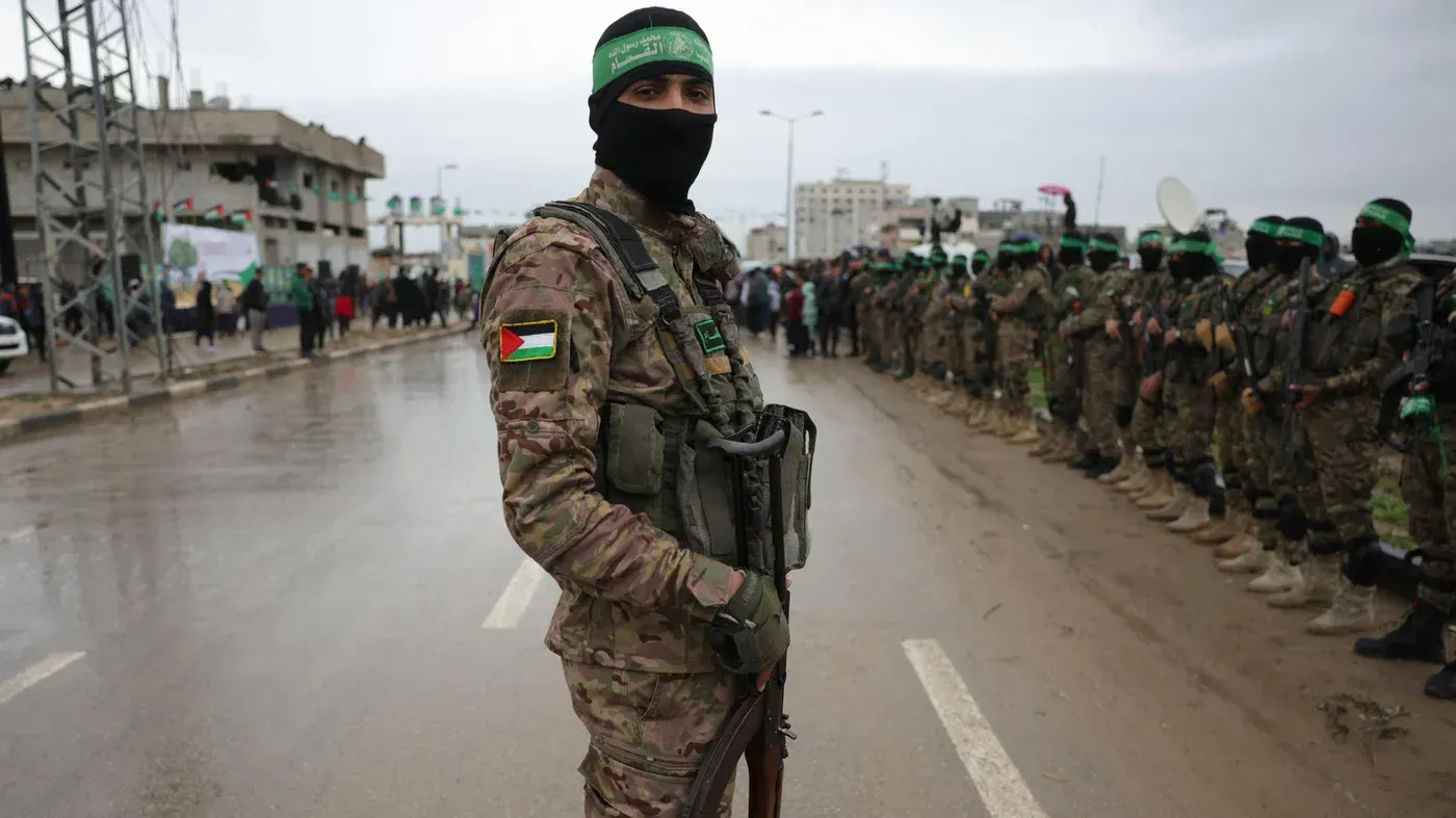 مقاتل من حماس يرتدي زيًا عسكريًا ويقف في مقدمة صف من الجنود خلال استعراض، مع العلم الفلسطيني على ذراعه.