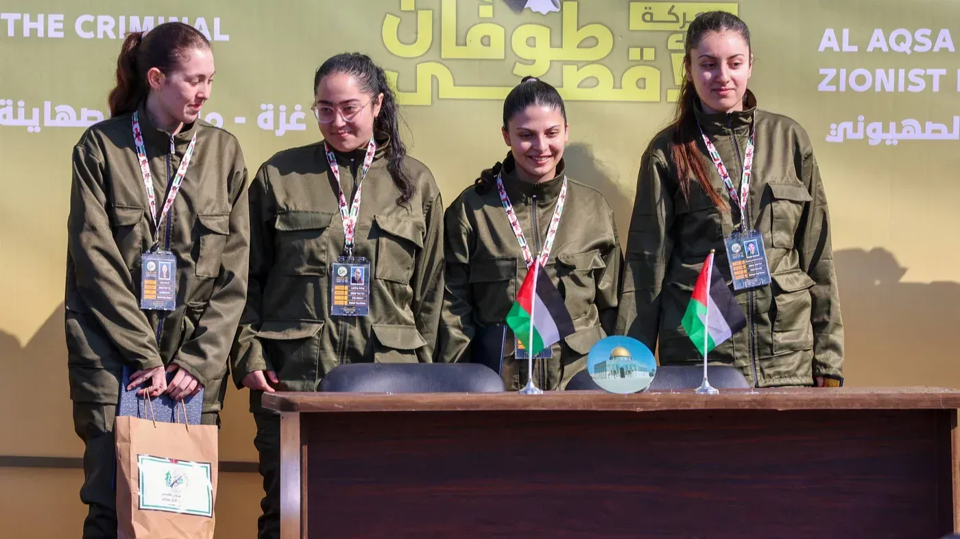 أربع مجندات إسرائيليات يرتدين زيًا عسكريًا، يقفن أمام منصة تحمل أعلامًا فلسطينية، وذلك بعد الإفراج عنهن كجزء من اتفاق هدنة.