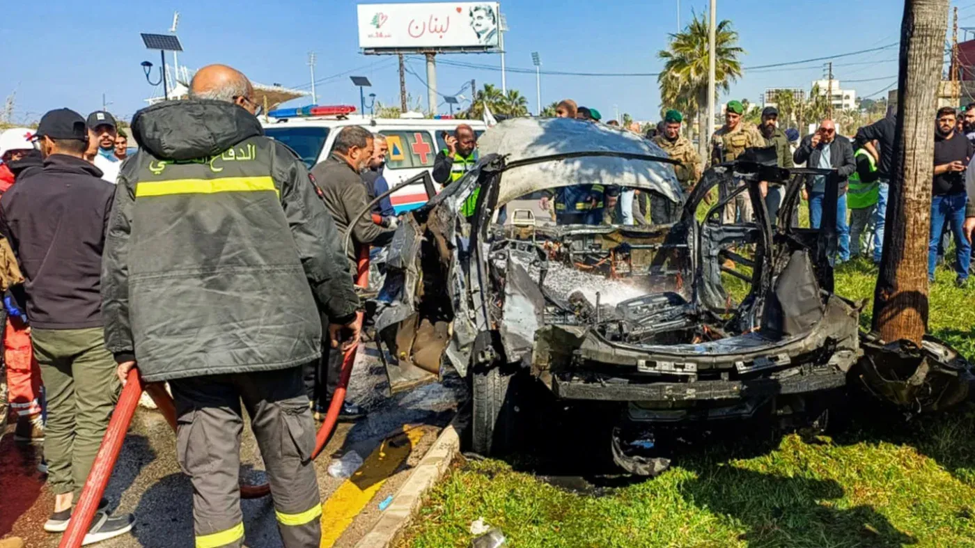 موقع حادث اغتيال محمد إبراهيم شاهين في صيدا، يظهر سيارة مدمرة بفعل الغارة الجوية، مع وجود رجال إطفاء ومارة في الخلفية.