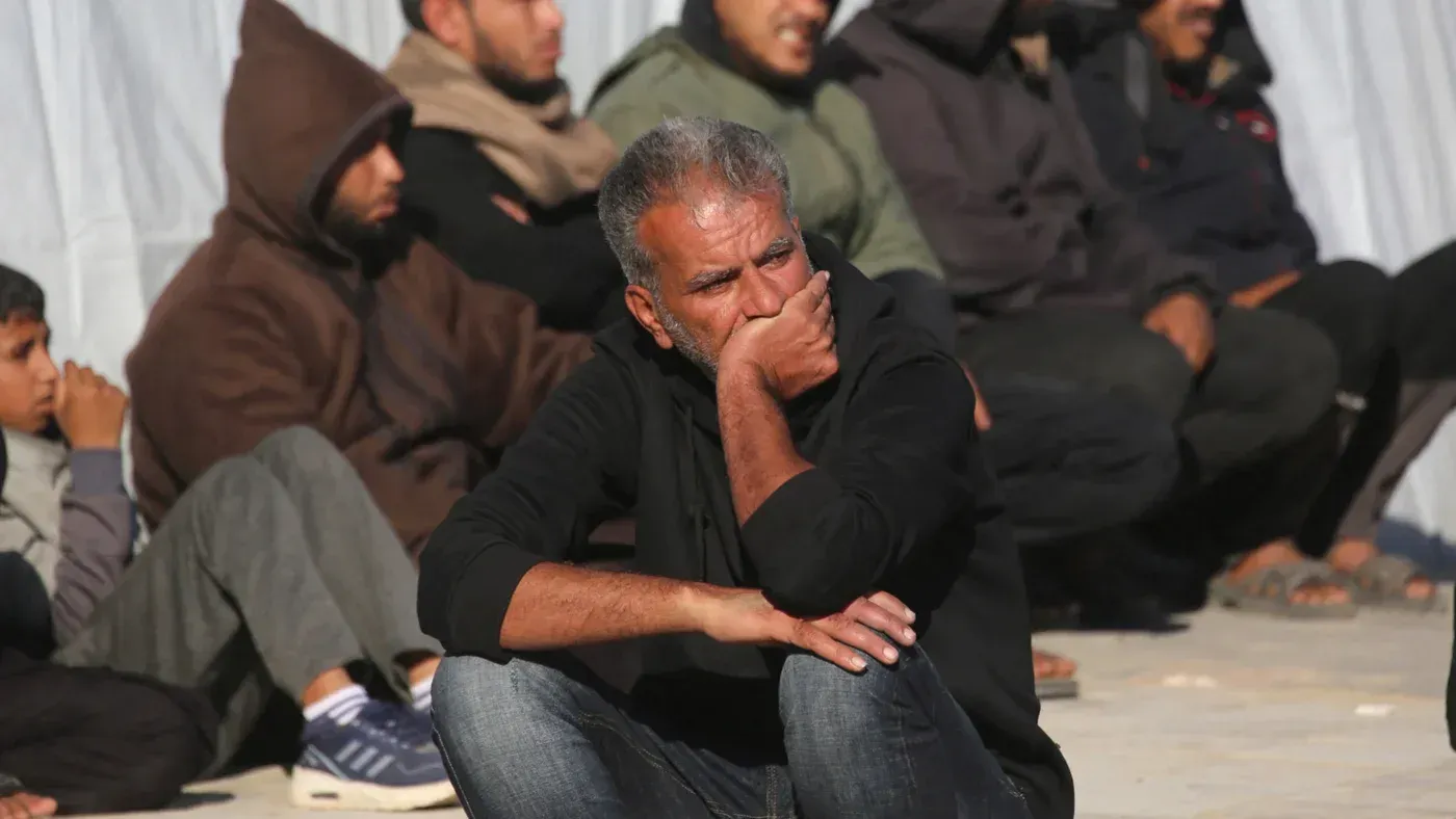 رجل يجلس على الأرض في غزة، يبدو عليه القلق والتفكير، بينما خلفه مجموعة من الرجال يجلسون في انتظار. تعكس الصورة التوتر والضغط في سياق المفاوضات السياسية.