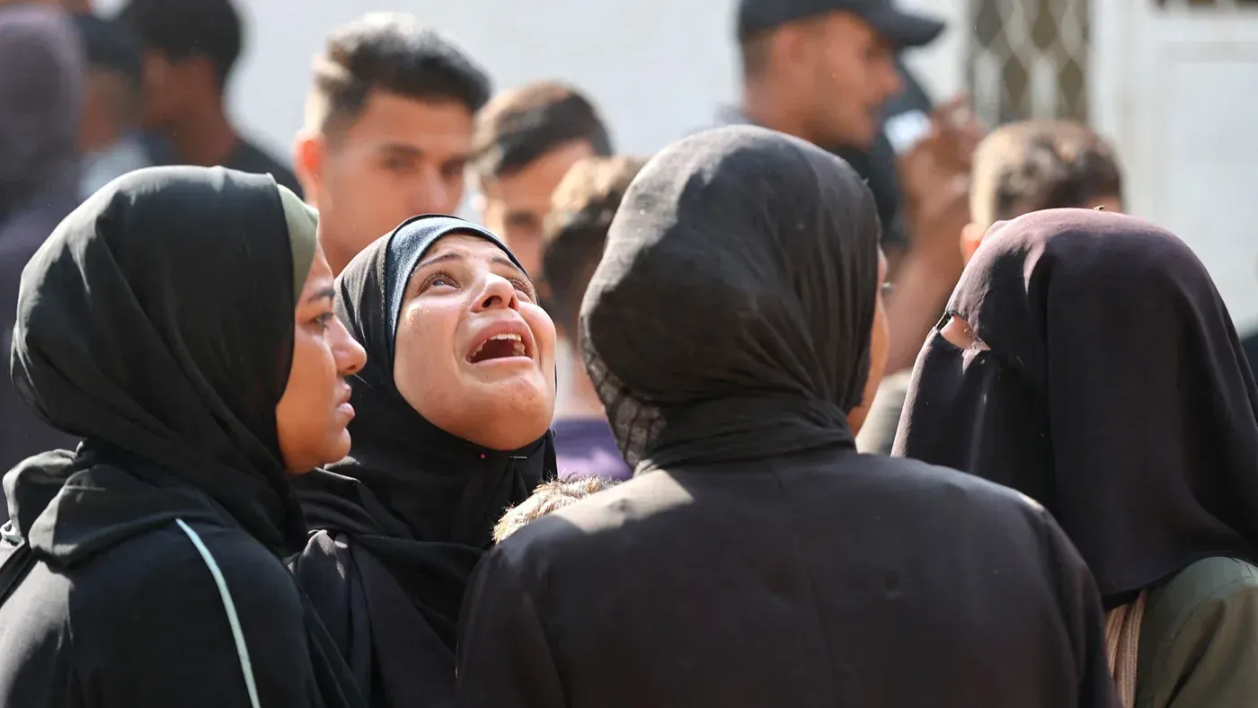 نساء يرتدين الحجاب في حالة من الحزن، يعبّرن عن مشاعر الألم بعد الهجمات الإسرائيلية على غزة، وسط تجمع حشود في الخلفية.