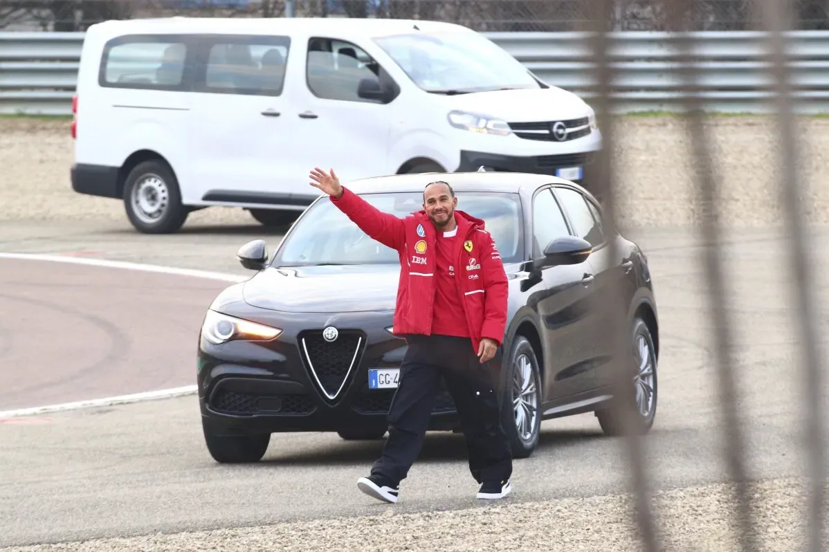 لويس هاميلتون يرتدي زي فيراري الأحمر، مبتسمًا ويلوح للمشجعين أثناء قيادته سيارة Alfa Romeo على حلبة فيورانو، مع حشود من المشجعين خلفه.