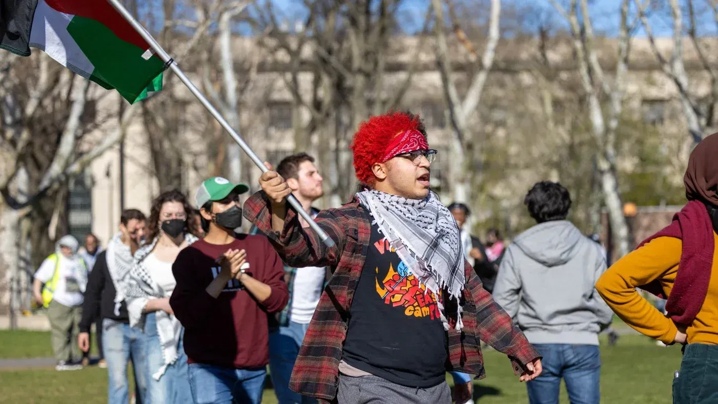 طالب يحمل علم فلسطين أثناء احتجاج في الحرم الجامعي، مع مجموعة من الطلاب في الخلفية، معبرًا عن دعم حقوق الفلسطينيين.