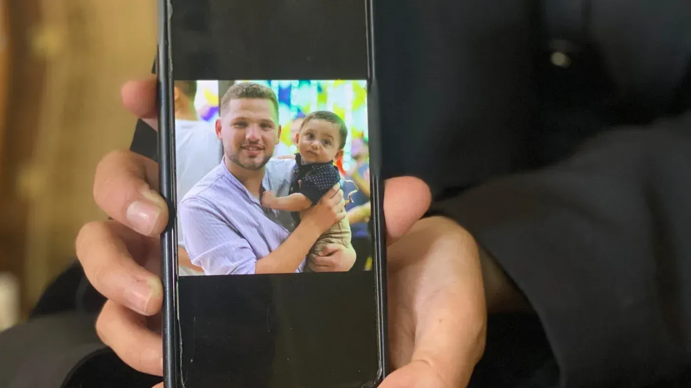 صورة تُظهر معاذ أبو ركبة، طبيب بيطري فلسطيني، يحمل طفلاً، تعبير عن الحب والعناية وسط الأوضاع الصعبة في غزة.