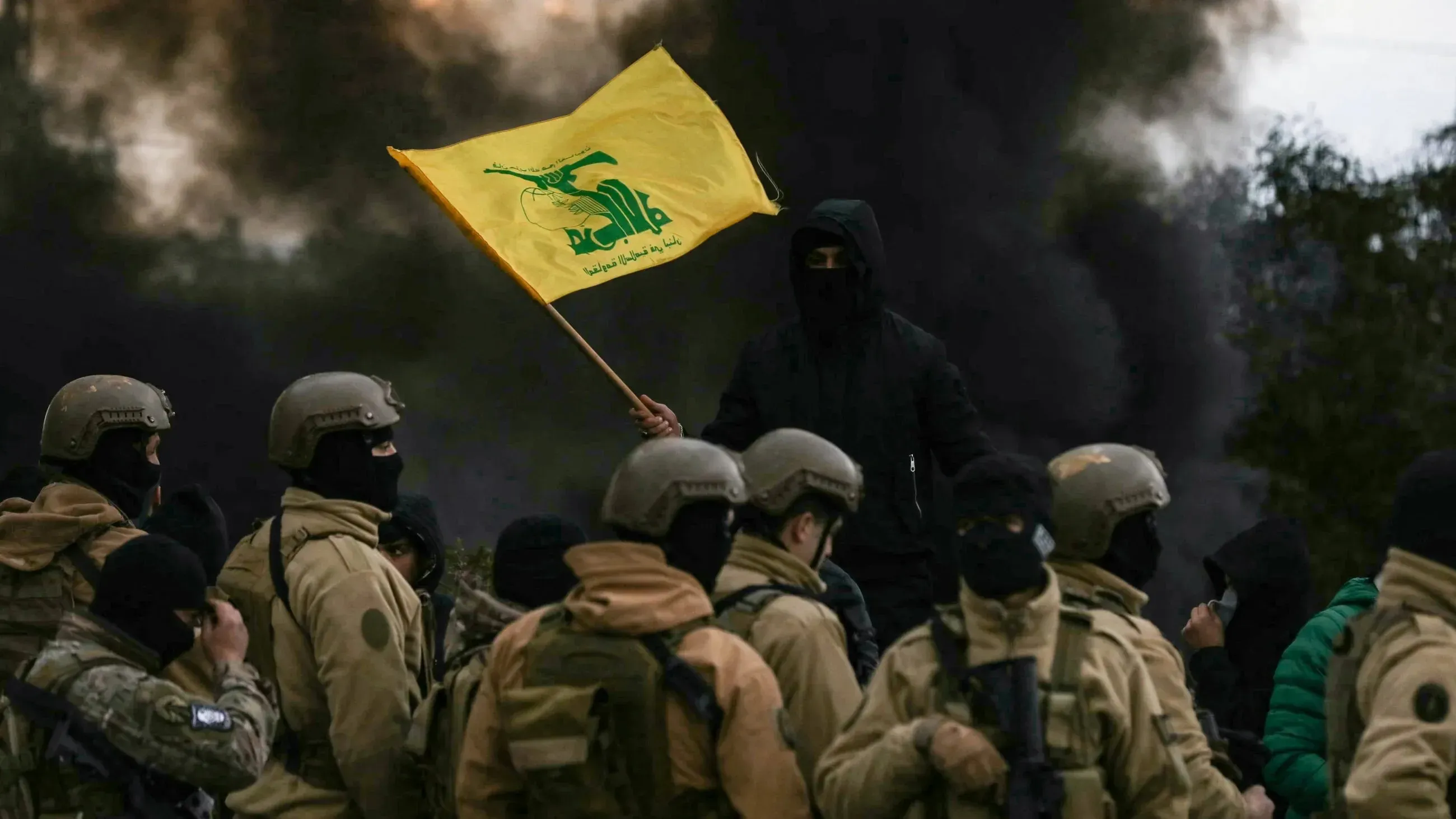 شخص يرتدي زي عسكري يحمل علم حزب الله الأصفر، محاطًا بجنود آخرين في موقع مظلم، مما يعكس التوترات السياسية في لبنان.
