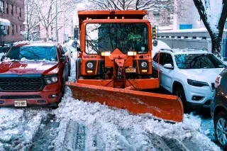 جرافة ثلوج برتقالية تعمل على إزالة الثلوج من شارع مزدحم بين سيارات متوقفة، مما يعكس جهود سيراكيوز في تحسين خدمات إزالة الثلوج.