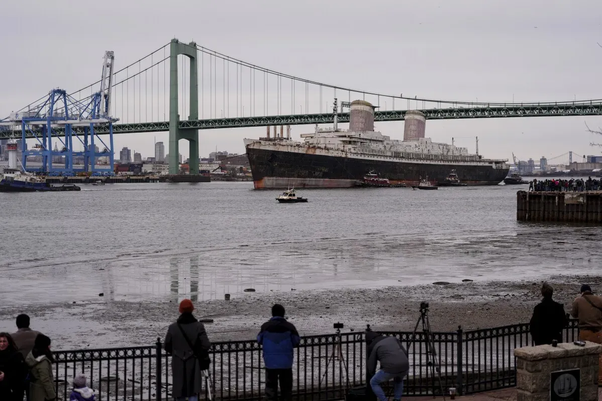 سفينة SS United States التاريخية تبحر عبر نهر ديلاوير، مع جسر خلفي ومشاهدين على الواجهة البحرية، استعدادًا لتحويلها لشعاب مرجانية اصطناعية.