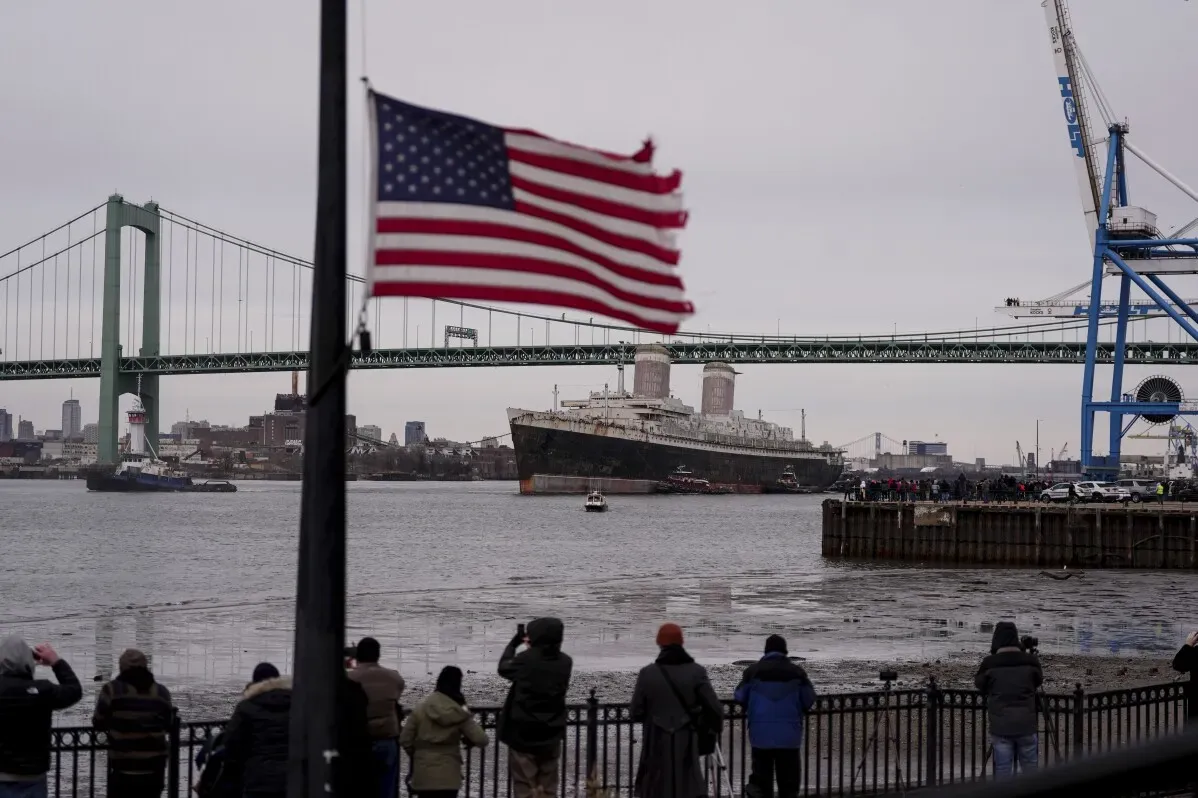 سفينة SS United States التاريخية تغادر نهر ديلاوير، مع جسر خلفي وعلم أمريكي، استعدادًا لتحويلها إلى شعاب مرجانية اصطناعية في فلوريدا.